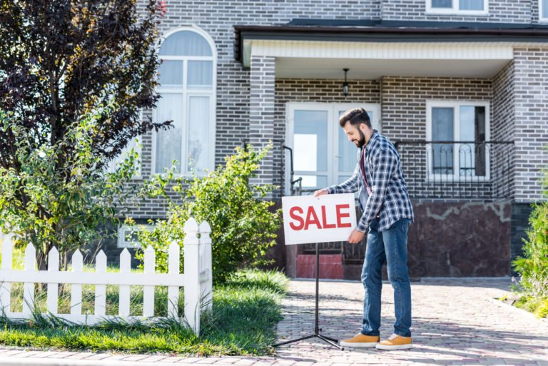Man holding "For Sale" sign in front of a house, real estate and home selling.